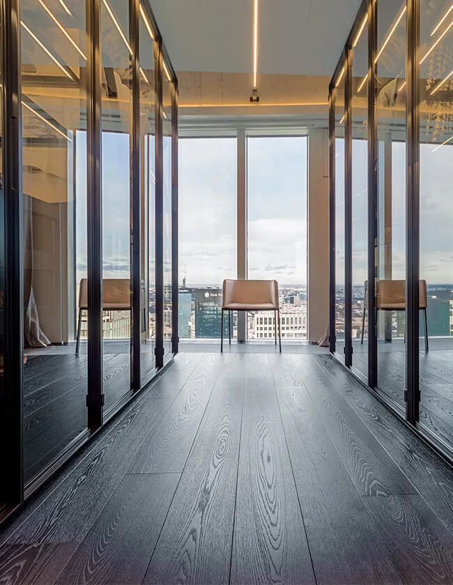 Pohled na tmavou dubovou podlahu v interiéru soukromého bytu Zlota ve Varšavě, kde výrazná kresba dřeva směřuje k prosklené stěně s výhledem na panorama města, lemovanou zrcadlovými plochami.