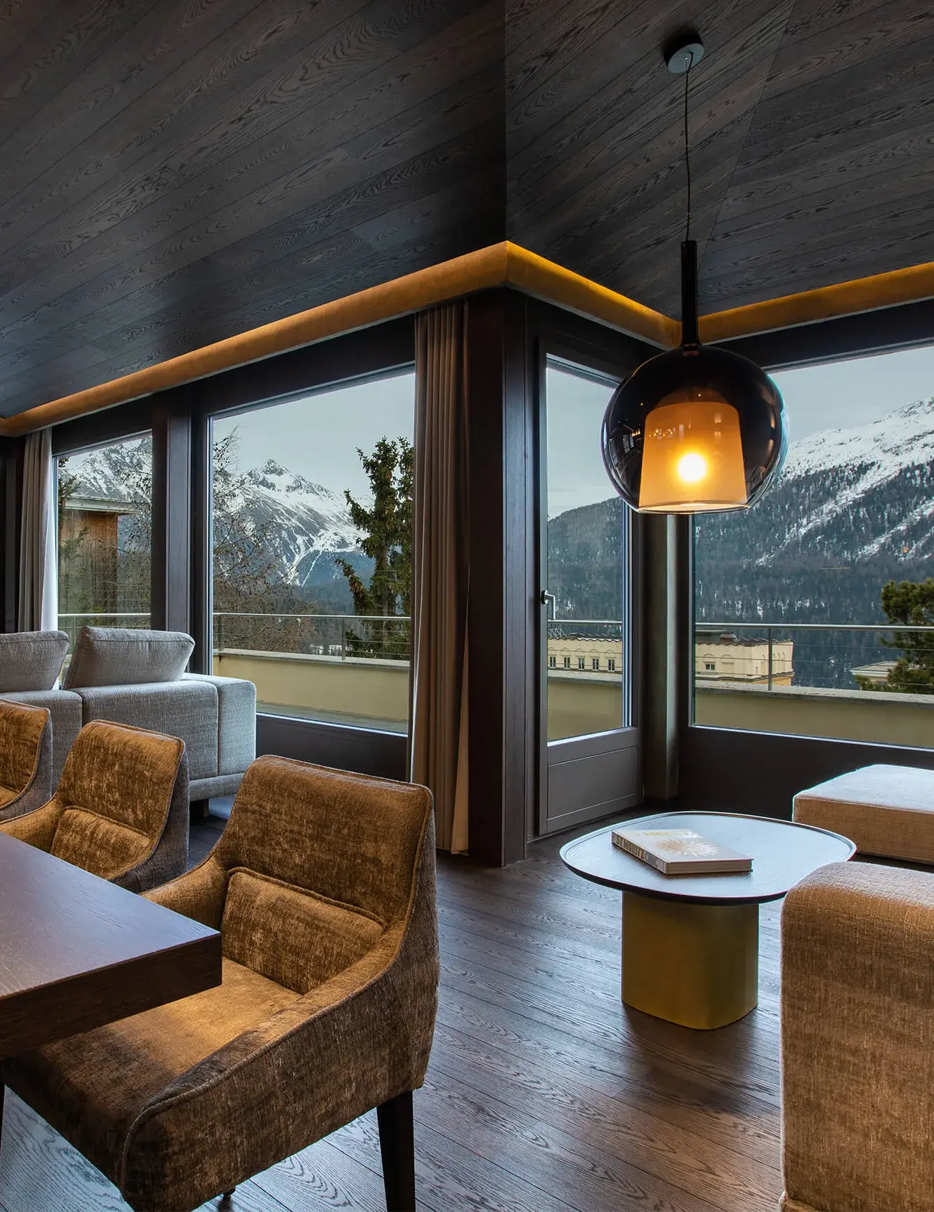 Luxusní interiér rezidence ve Svatém Mořici s tmavou dubovou podlahou a ladícím dřevěným podhledem, které doplňuje elegantní nábytek v hnědých tónech a panoramatický výhled na zasněžené vrcholky Alp skrze velkoformátová okna.