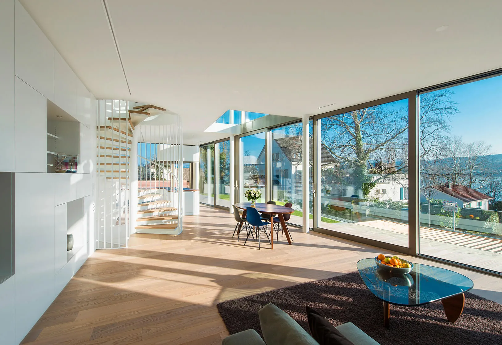 Prosvětlený interiér vily Flexhouse u Curyšského jezera se světlou dubovou podlahou, kterému dominuje bílé točité schodiště a prosklené stěny nabízející nerušený výhled do zahrady a na hladinu jezera.