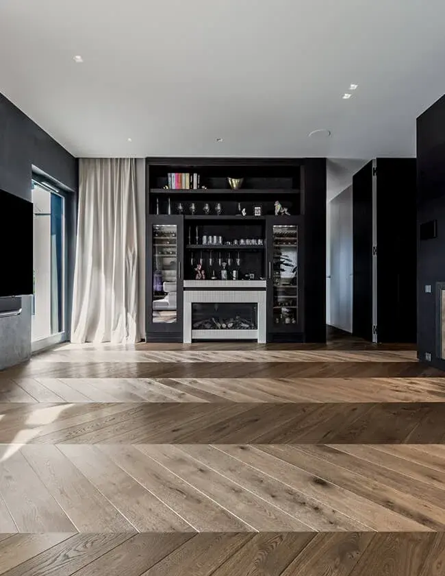 Moderní interiér obývacího pokoje v soukromém bytě Skierniewicka s výraznou dubovou podlahou položenou ve vzoru maďarského stromečku (chevron), která kombinuje světlé a tmavé odstíny dřeva a doplňuje tmavý vestavěný nábytek s krbem.