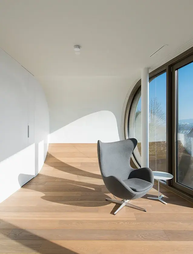 Minimalistický interiér Flex House v Curychu s ikonickým šedým křeslem Egg, kde prohnutá bílá stěna plynule přechází v prosklenou plochu s výhledem na Curyšské jezero a prosvětluje kartáčovanou dubovou podlahu