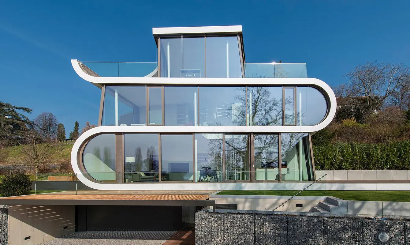 Exteriér vily Flexhouse v Curychu s unikátní bílou fasádou ve tvaru stuhy a velkoformátovým prosklením, která svou dynamickou architekturou odráží pohyb okolní krajiny, jezera a blízké železniční trati.