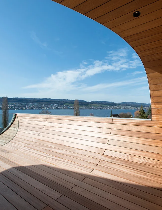 Detailní pohled z horní terasy vily Flexhouse na Curyšské jezero, kde zaoblené dřevěné obložení fasády plynule přechází v podlahu terasy a rámuje panoramatický výhled na protější břeh.