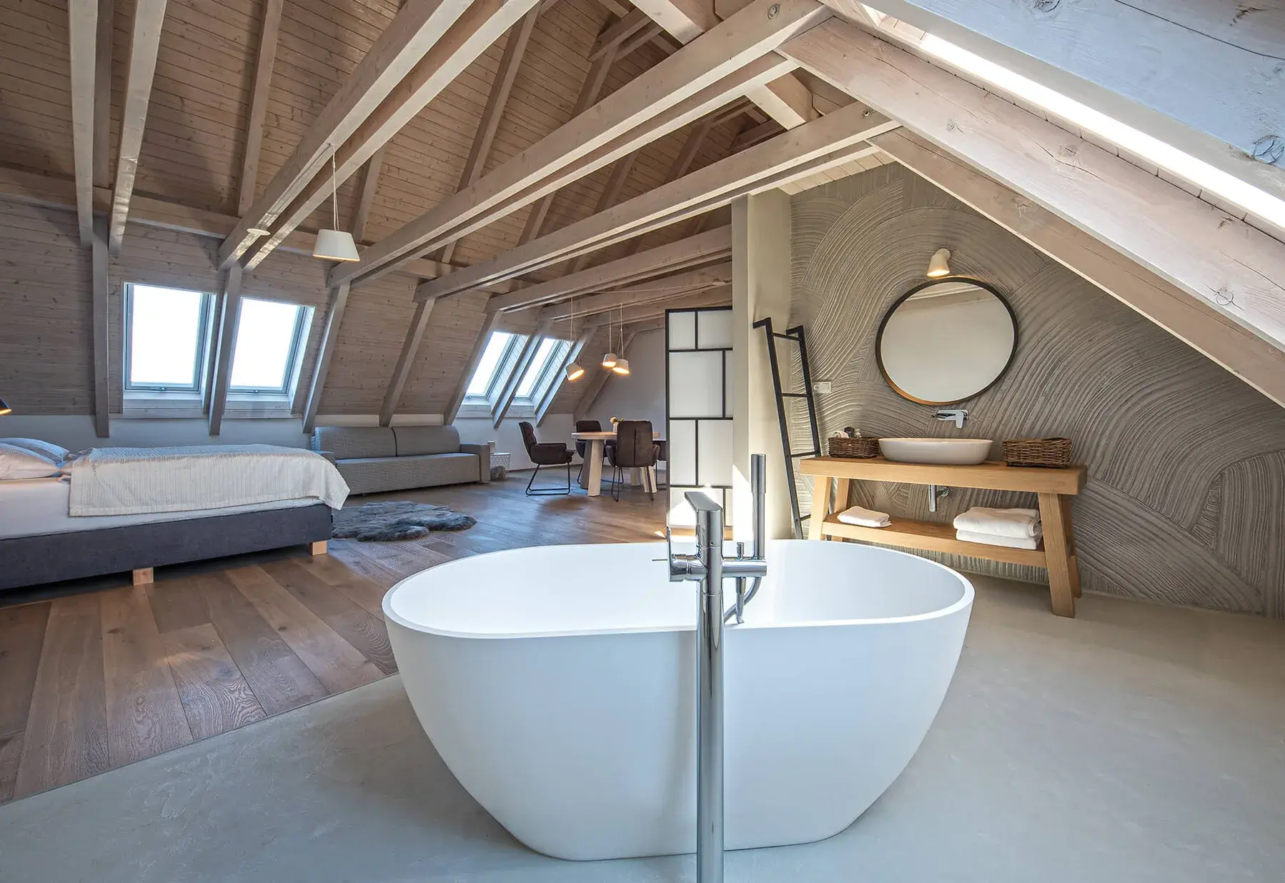 Moderní otevřený prostor podkrovního apartmánu s přiznanými trámy v šedém dekoru Smoke, kde dřevěná podlaha v ložnici plynule přechází v litou stěrku v koupelně s volně stojící vanou a designovou stěnou se strukturovanou omítkou.