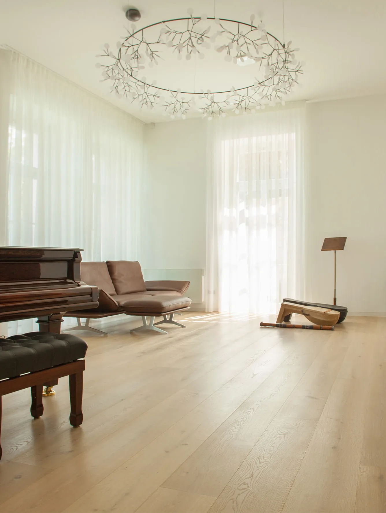 Prosvětlený obývací pokoj s přírodní dubovou podlahou, hnědou koženou pohovkou a klavírem, kterému dominuje velké designové kruhové svítidlo a jemné bílé závěsy.