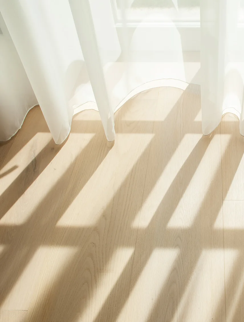 Detailní záběr na prosvětlenou dubovou podlahu, kde sluneční paprsky pronikající skrze jemné bílé závěsy vytvářejí hru světla a stínu na přírodní textuře dřeva.