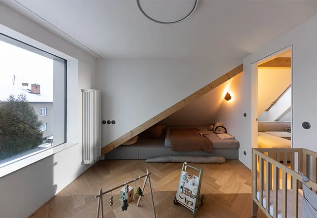 Moderní podkrovní dětský pokoj s dubovou podlahou položenou do vzoru stromečku, který doplňuje atypické lůžko vestavěné pod šikminou střechy s hřejivým LED osvětlením a výhledem do zasněžené krajiny velkým oknem.
