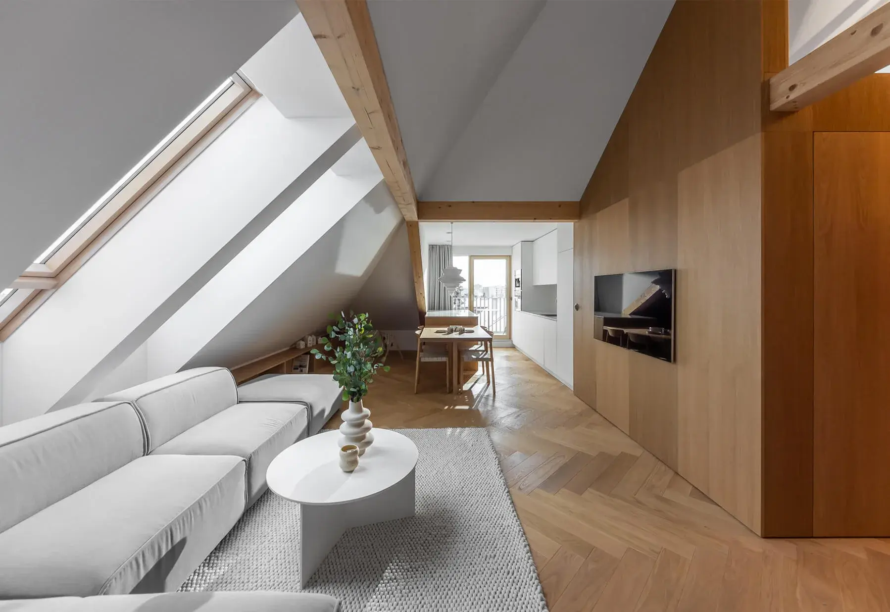 Moderní podkrovní obývací pokoj s dubovou podlahou položenou do vzoru stromečku, který plynule navazuje na celodřevěné obložení stěny s integrovanou TV, doplněný o světlou pohovku a konferenční stolek pod velkorysými střešními okny.