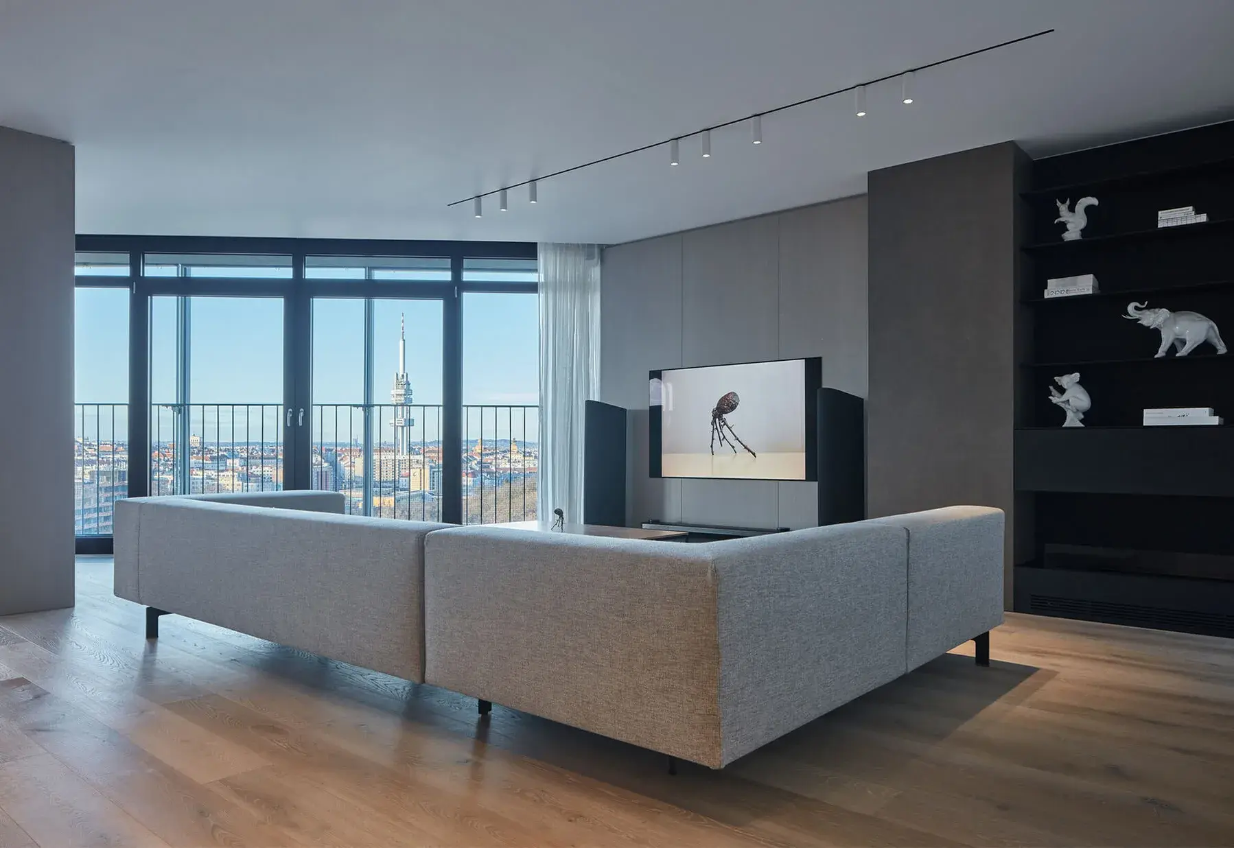 Moderní minimalistický interiér obývacího pokoje s velkou šedou rohovou pohovkou a dřevěnou podlahou z kolekce Heritage, s výhledem na panorama města s dominantou Žižkovské televizní věže.