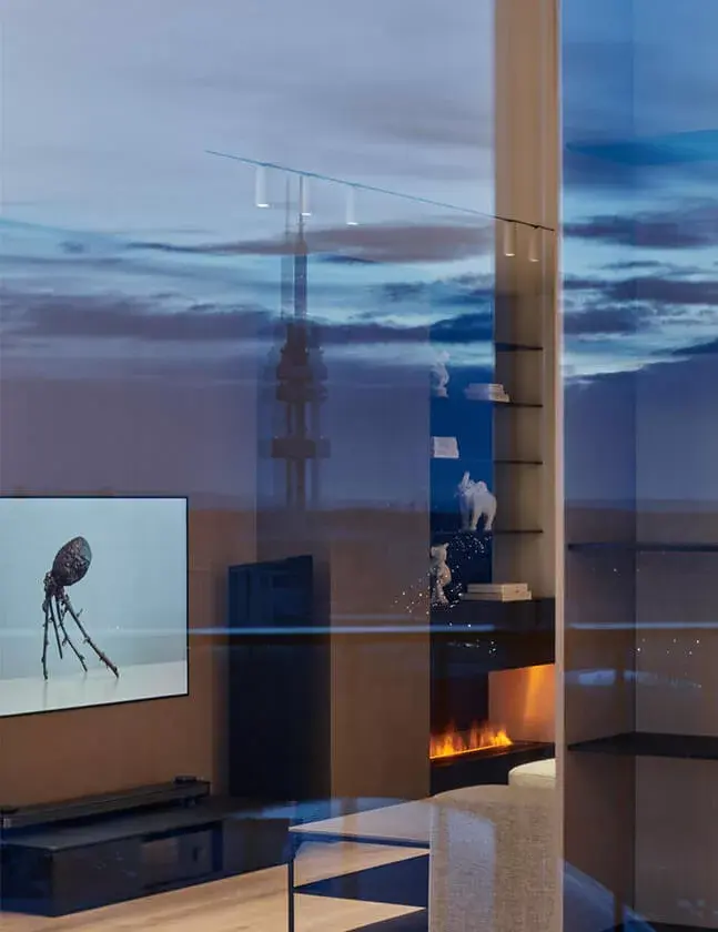 Večerní pohled na moderní interiér bytu s krbem, v jehož oknech se zrcadlí panorama Prahy s osvětlenou Žižkovskou televizní věží pod dramatickou soumračnou oblohou.