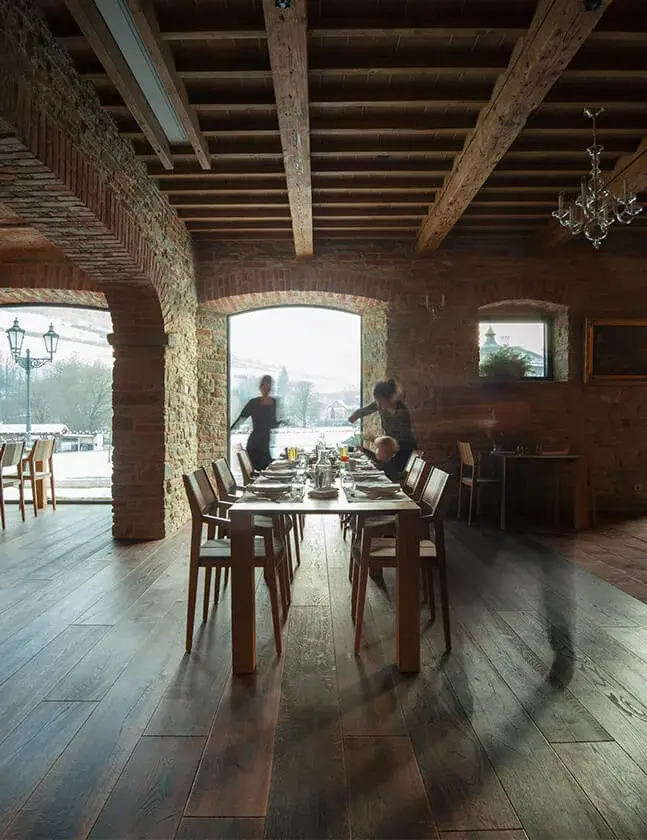 Rustikální jídelna ve vile Bianca s tmavou dubovou podlahou Oakcent a masivním trámovým stropem, zasazená do historického prostoru s odhaleným cihlovým a kamenným zdivem, s výhledem do zasněžené zahrady.