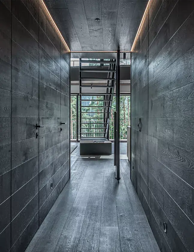 Moderní chodba v Apartmánu Lipno s celodřevěným obložením stěn a stropu, které plynule navazuje na tmavou dubovou podlahu Oakcent a směřuje k industriálnímu schodišti s výhledem do šumavské přírody.