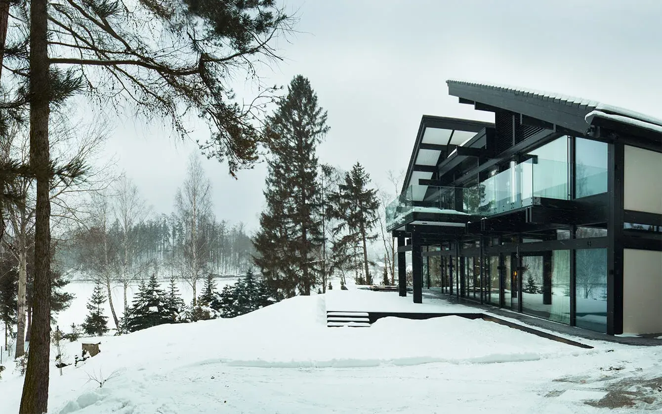 Moderní černá prosklená architektura Vily Trnávka citlivě zasazená do zasněžené zimní krajiny u břehu jezera, obklopená vzrostlými jehličnany a břízami.