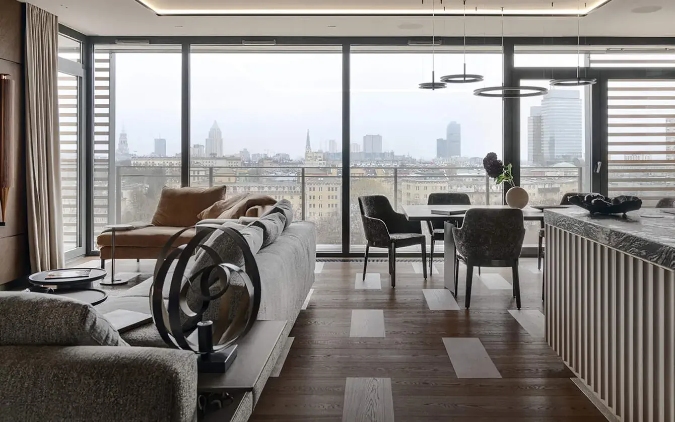 Panoramatický pohled na obývací prostor bytu Grzybowska ve Varšavě s prosklenou stěnou odhalující výhled na město, kde tmavá dubová podlaha s pravidelně vloženými světlými poli vizuálně propojuje relaxační zónu a jídelnu.
