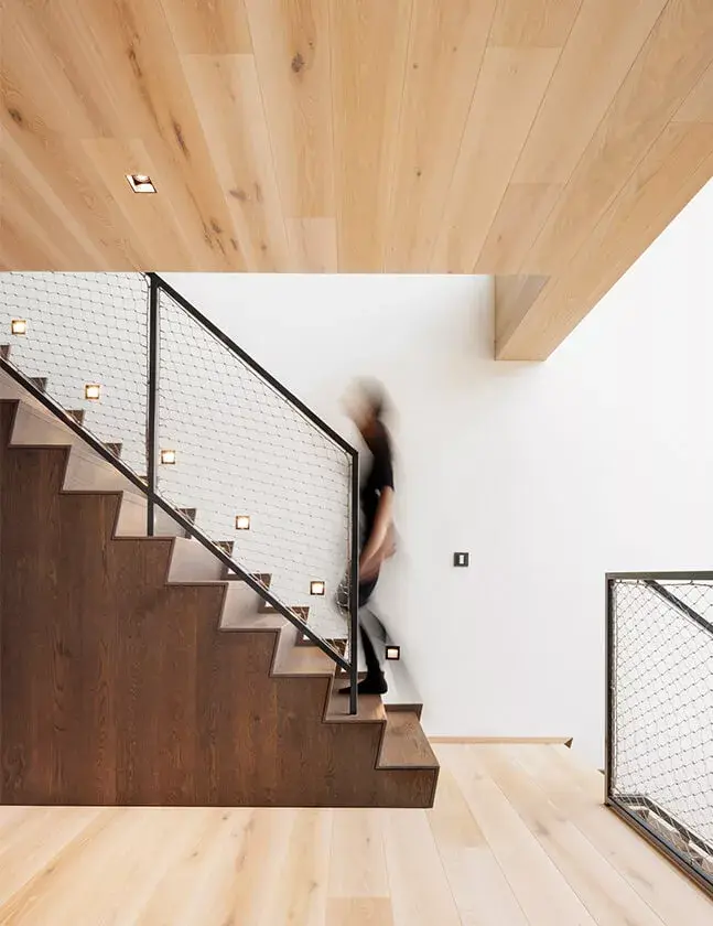 Moderní schodiště v soukromém domě v Židlochovicích s nášlapy z tmavého dubu, které kontrastují se světlou dubovou podlahou a dřevěným podhledem, doplněné o minimalistické síťované zábradlí s integrovaným bodovým osvětlením.