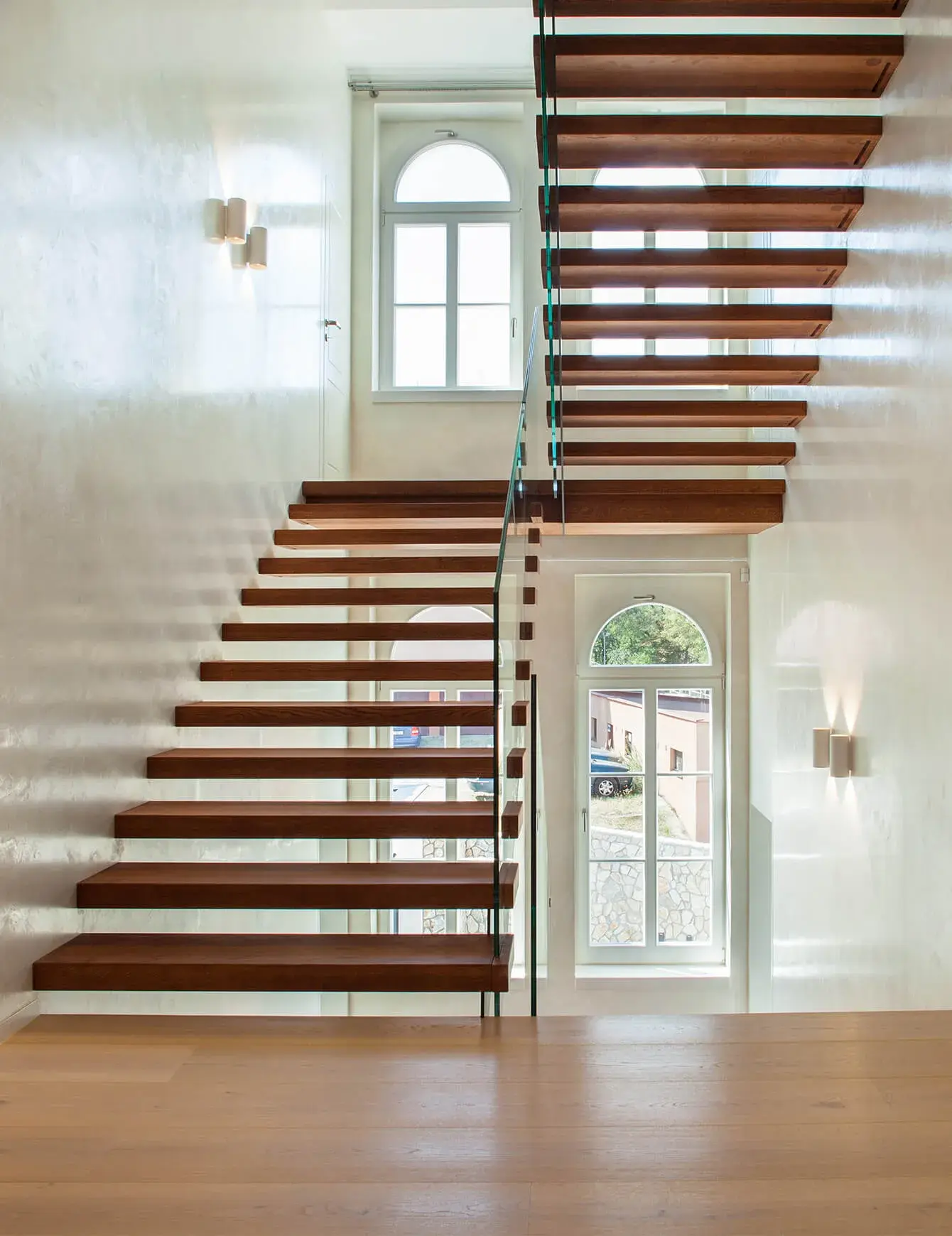 Moderní samonosné dřevěné schodiště ve vile Blaženka s masivními dubovými stupni a celoskleněným zábradlím, které v kombinaci se světlou stěrkou na stěnách a klenutými okny propouští do interiéru dostatek přirozeného světla.