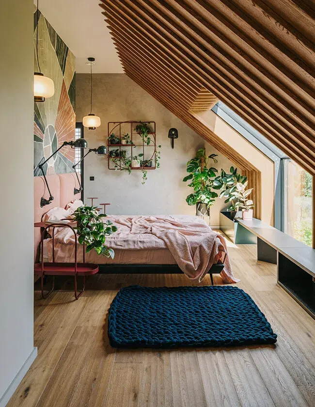 Útulná podkrovní ložnice v projektu Stodola v Miloslavi s přírodní dubovou podlahou a výrazným lamelovým podhledem, který sleduje šikminu střechy a doplňuje moderní interiér s růžovými akcenty a množstvím pokojových rostlin.