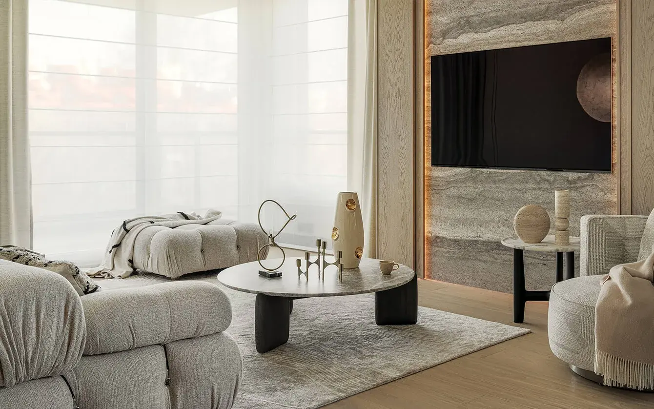 Prosvětlený interiér obývacího pokoje v soukromém bytě Iwicka ve Varšavě s jemnou dubovou podlahou, kterou doplňuje kamenný obklad stěny s integrovaným podsvícením, mramorové konferenční stolky a designový nábytek v neutrálních krémových tónech.