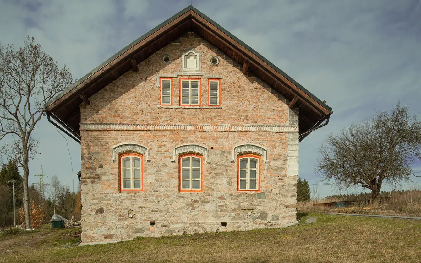 Pohled na štít historické Chalupy U Cesty s tradiční fasádou kombinující odhalené kamenné zdivo a režné cihly, doplněný o dřevěné podbití střechy a původní členěná okna s výrazným olemováním.