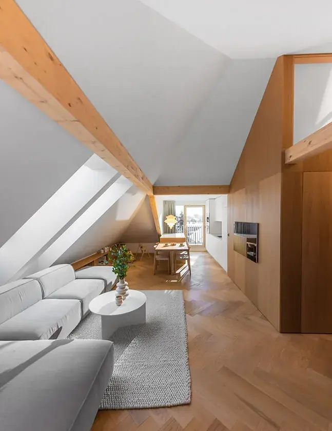Světlý podkrovní obývací pokoj ve střešním bytě v Olomouci s masivními přiznanými trámy a dubovou podlahou položenou do vzoru stromečku, který doplňuje moderní šedá sedací souprava a dřevěné vestavné skříně kopírující sklon střechy.