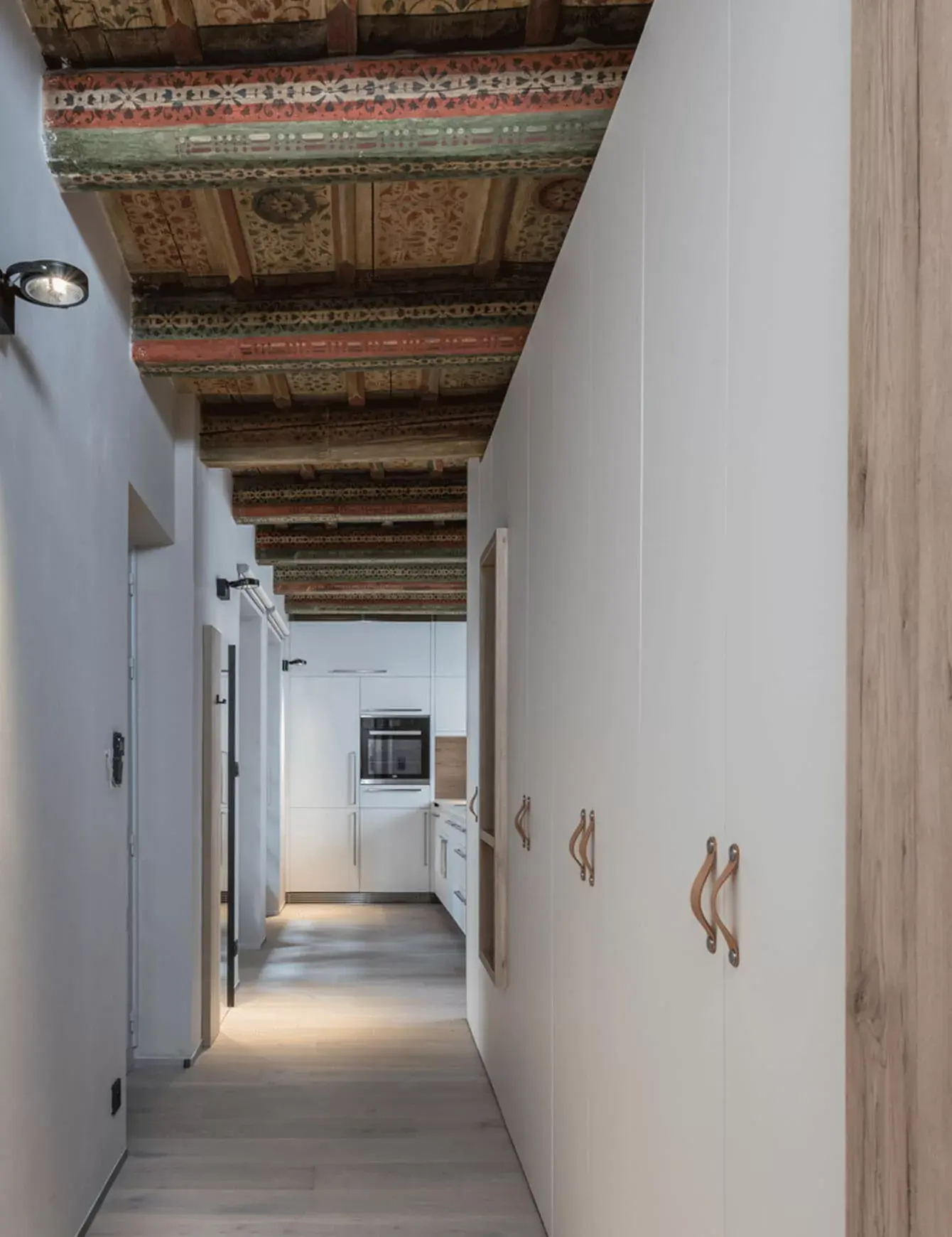 Moderní minimalistická chodba v historickém Bytě na Malé Straně s bílými vestavnými skříněmi a světlou dubovou podlahou, kterou zdobí restaurovaný renesanční trámový strop s bohatou dekorativní malbou.