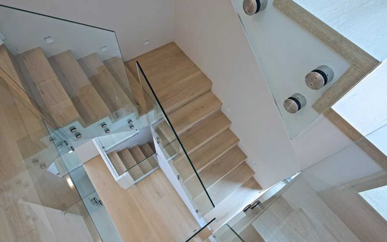 Pohled shora na moderní dřevěné schodiště v soukromém domě na Moravě se světlými dubovými stupni a minimalistickým celoskleněným zábradlím s nerezovými úchyty, které doplňuje světlou dřevěnou podlahu v hale.