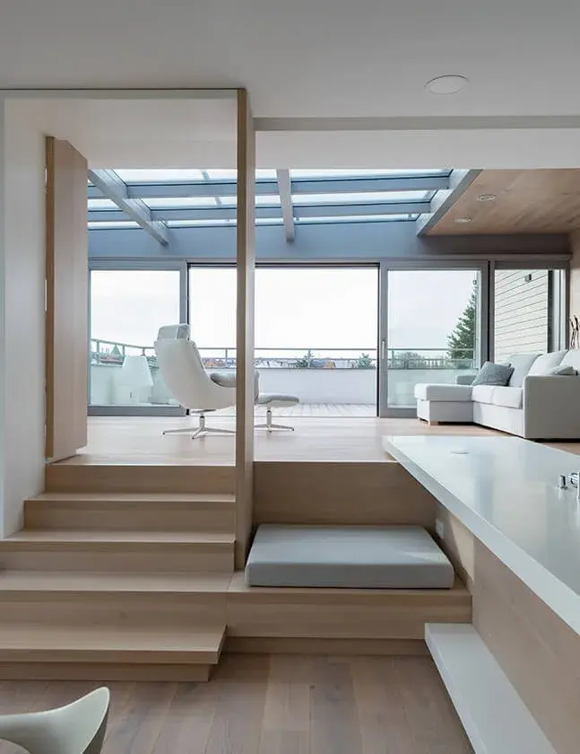 Moderní prosvětlený interiér Střešního bytu s víceúrovňovou dřevěnou podlahou, kde schodišťové stupně plynule přecházejí v relaxační pódium pod prosklenou střechou terasy, doplněný minimalistickým bílým nábytkem a designovým křeslem.