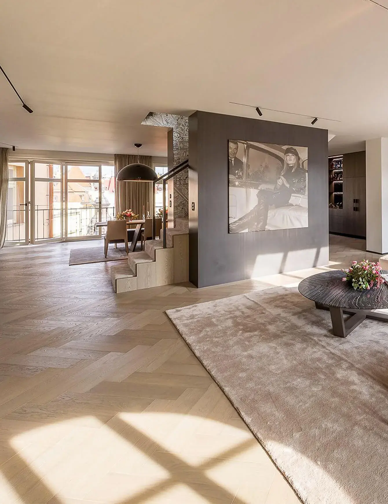 Luxusní interiér Soukromého bytu v Praze se světlou dubovou podlahou položenou do vzoru stromečku, kterému dominuje tmavá dělící stěna s uměleckou fotografií, doplňující otevřený prostor s jídelním koutem a velkorysým výhledem na město.