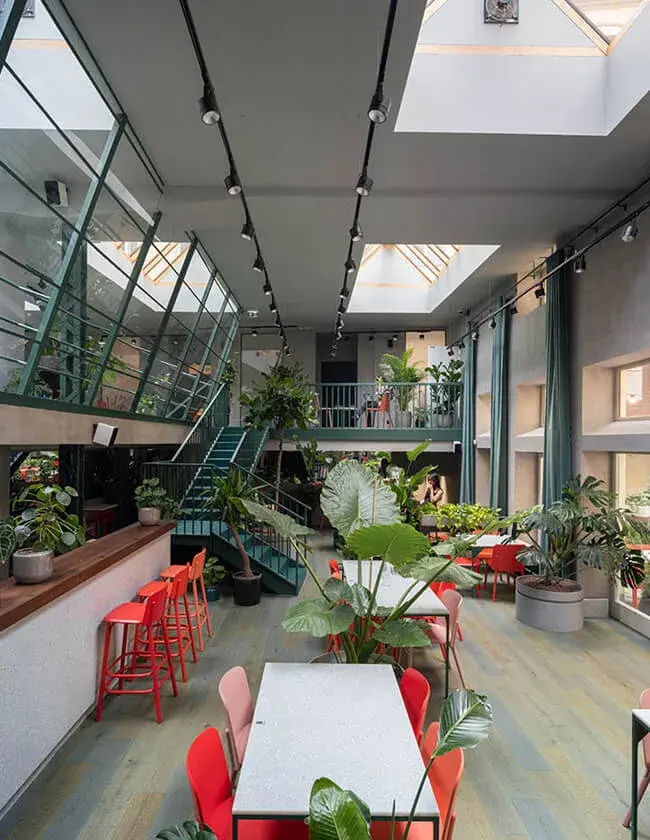 Moderní prosvětlený interiér Healthy Longevity Café s vysokými stropy a střešními okny, kterému dominuje množství živé zeleně, barevný nábytek v odstínech červené a růžové a výrazné zelené schodiště vedoucí na galerii.
