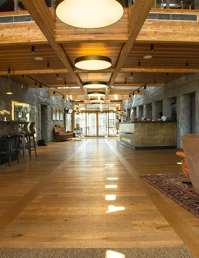 Rustikální interiér recepce Panorama Golf Resortu s kartáčovanou dubovou podlahou a masivním trámovým stropem s integrovaným kruhovým osvětlením, který doplňují stěny z nahrubo opracovaného kamene a elegantní barový pult s mosazným detailem.