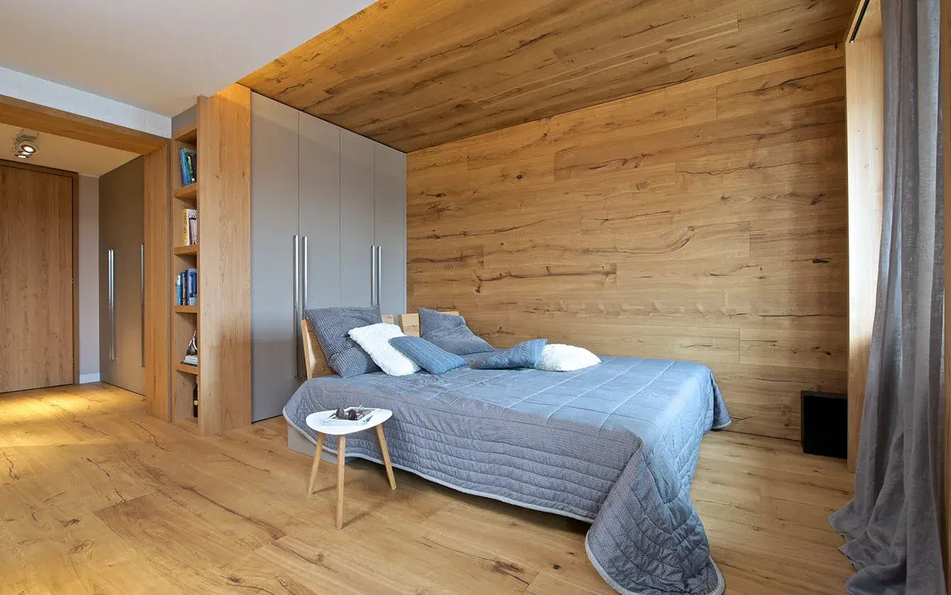 Moderní ložnice ve Valašském Apartmánu s celodřevěným konceptem, kde přírodní dubová podlaha plynule přechází v obklad stěn a stropu, doplněná o vestavné šedé skříně a minimalistické vybavení v přírodních tónech.