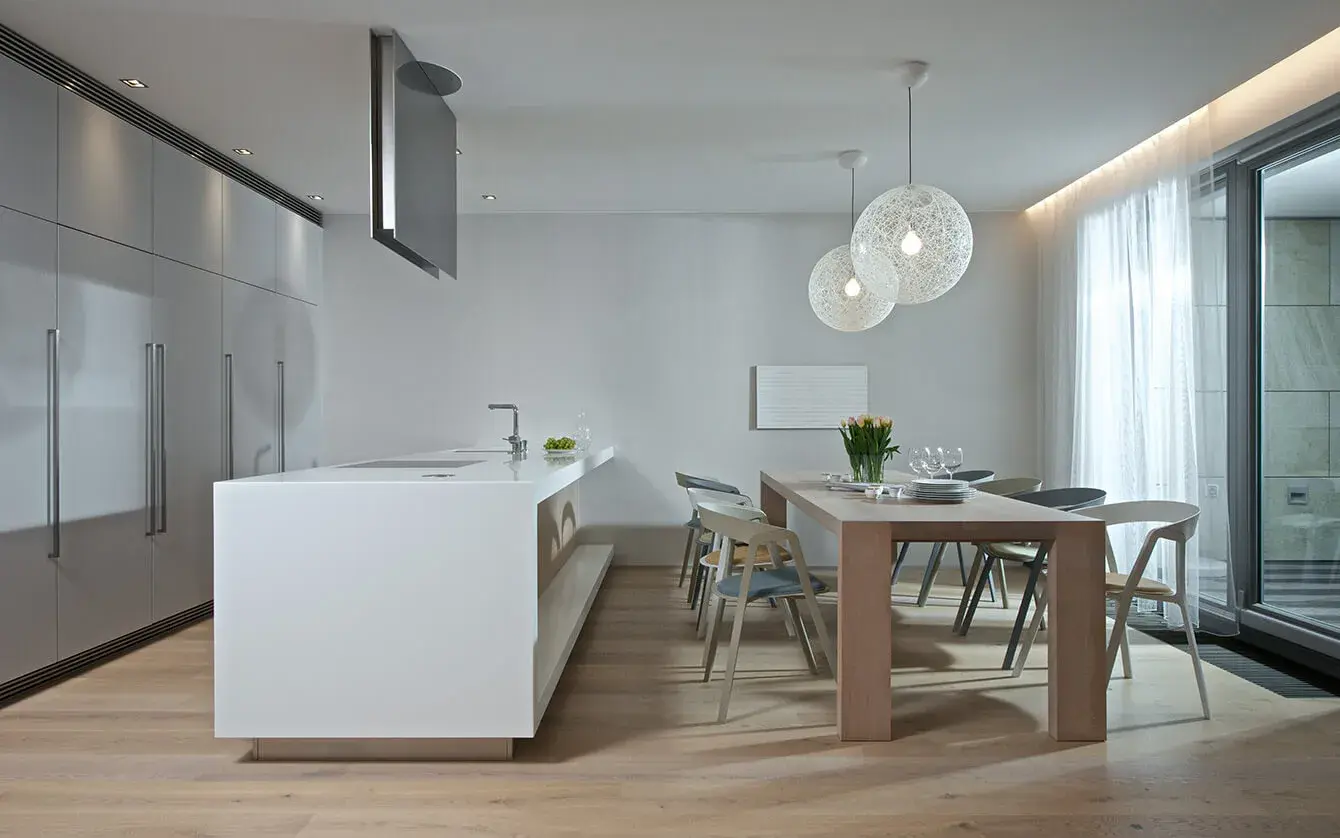 Moderní minimalistická kuchyně spojená s jídelnou v Soukromém bytě na nábřeží, kde světlá dubová podlaha harmonizuje s čistě bílým kuchyňským ostrovem, dřevěným jídelním stolem a designovými kulatými svítidly nad ním.