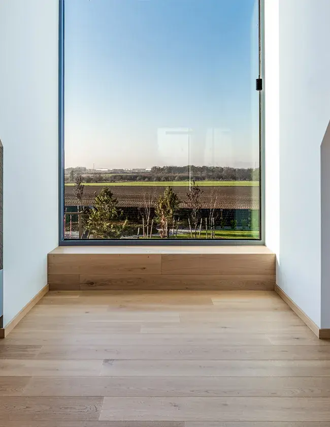 Moderní minimalistický interiér v Soukromém domě Tyniec Mały se světlou dubovou podlahou a vestavěným dřevěným sezením v okenním výklenku, který rámuje široký panoramatický výhled do okolní krajiny.