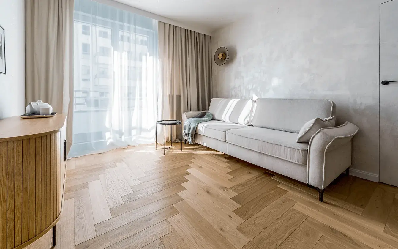 Prosvětlený interiér projektu Mini apartmán Varšava s klasickou dubovou podlahou položenou do vzoru stromečku, kterou doplňuje světlá pohovka, jemná dekorativní stěrka na stěnách a krémové závěsy propouštějící denní světlo.