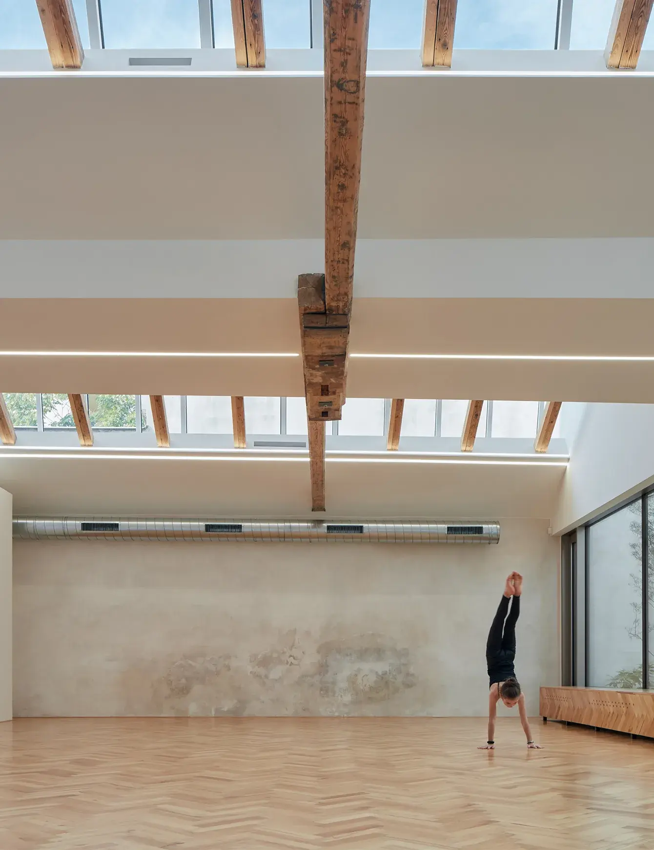 Moderní a vzdušný sál Yoga Garden & Art Gallery v Brně s přírodní dubovou podlahou položenou do vzoru stromečku, kterému dominují přiznané historické dřevěné trámy pod prosklenou střechou s lineárním LED osvětlením a minimalistická stěna s jemnou patinou.