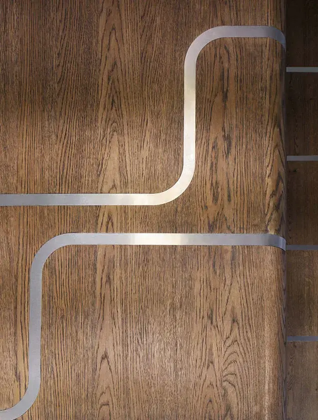 Detailní pohled na texturu kartáčovaného dubového dřeva Oakcent s integrovanou nerezovou lištou, která vytváří čistý přechod mezi přírodním materiálem a moderním kovem v interiéru Jetlag Baru.