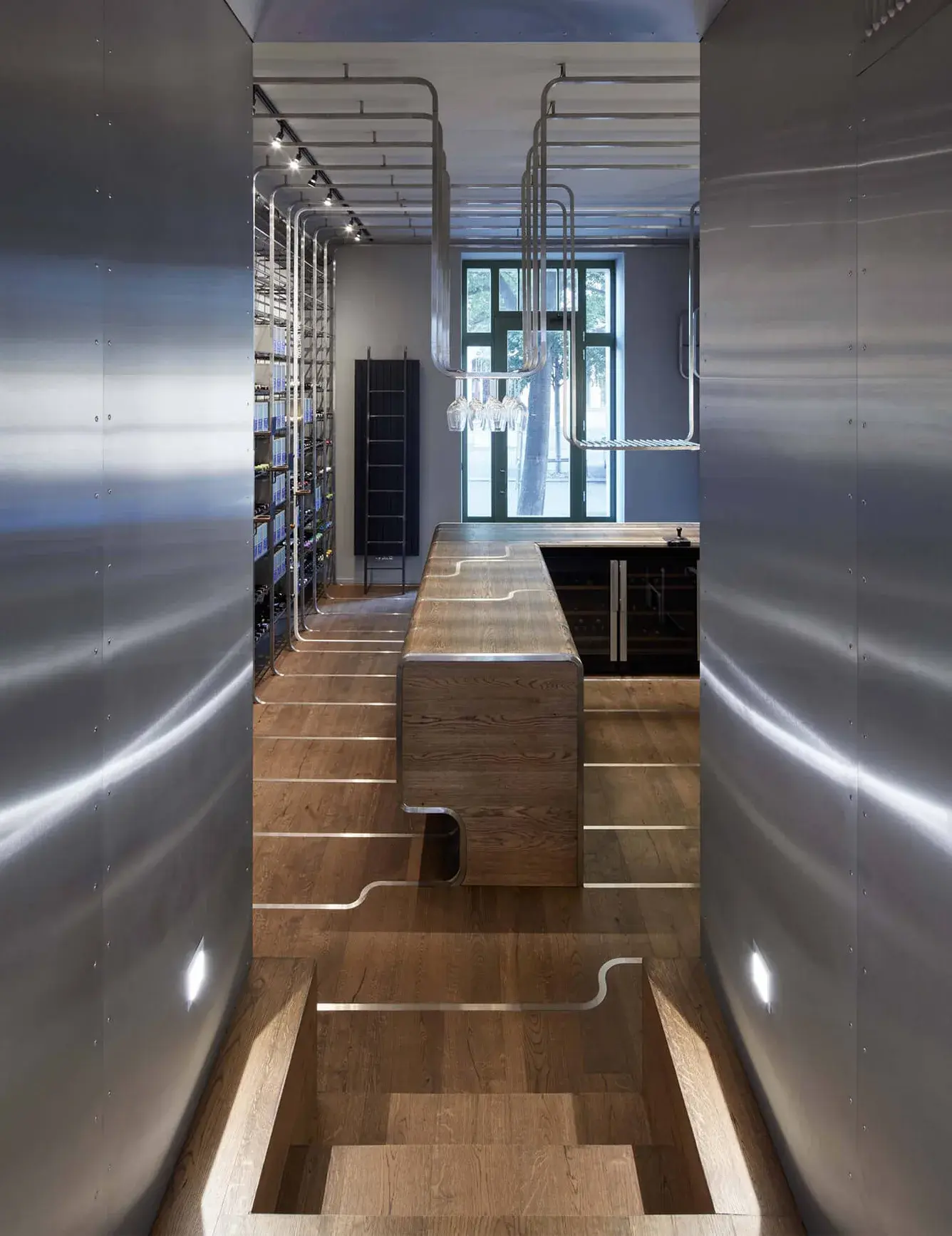 Industriální interiér Jetlag Baru v Praze s pohledem skrze nerezové obložení stěn na centrální dubový bar Oakcent s integrovanými kovovými lištami, které plynule přecházejí z podlahy na těleso pultu a doplňují geometrické závěsné systémy.