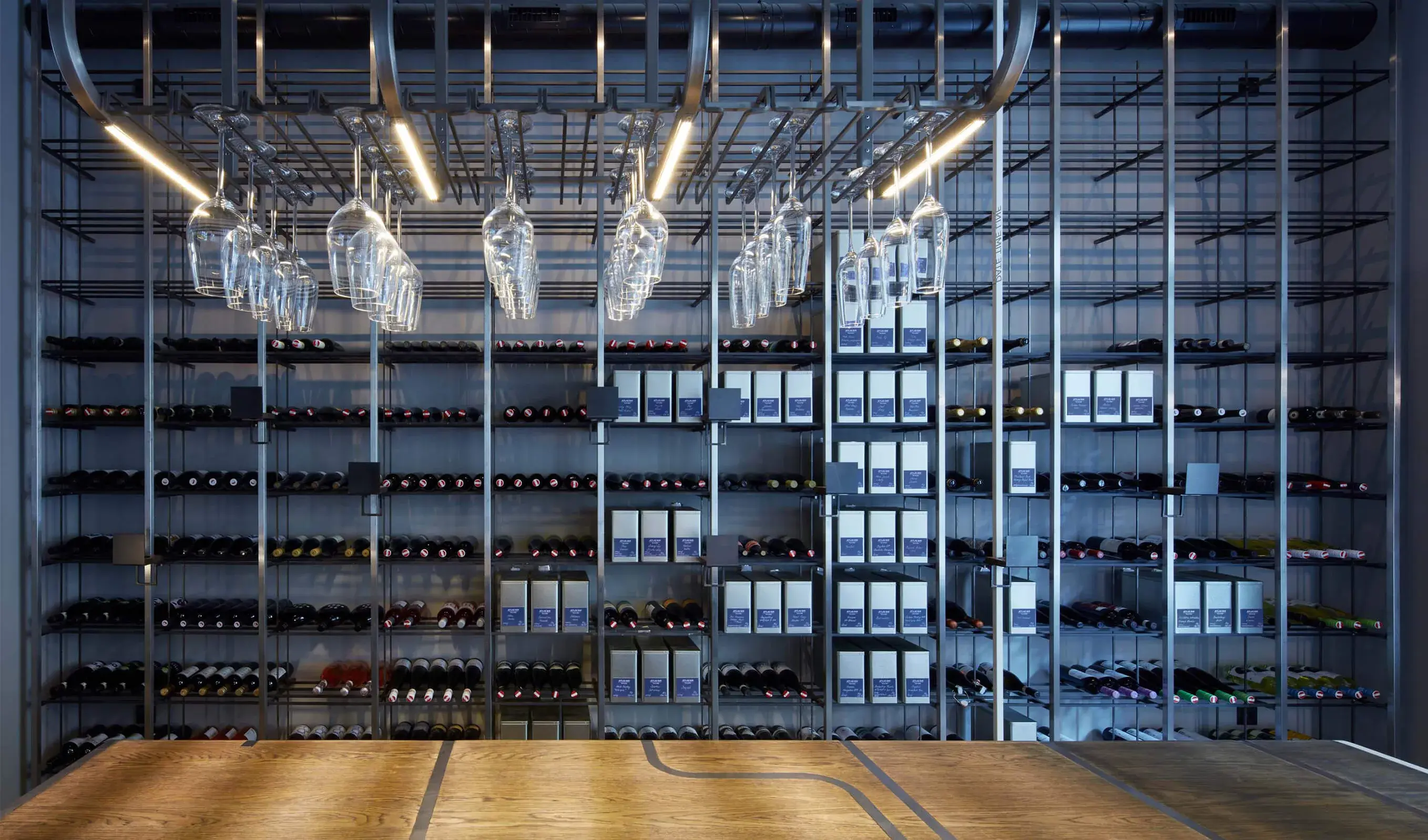 Pohled na prosvětlený regálový systém na víno v Jetlag Baru, který kombinuje kovovou konstrukci s dubovou odkládací plochou Oakcent Hazel s integrovanou nerezovou lištou a závěsným systémem pro vinné sklenice.