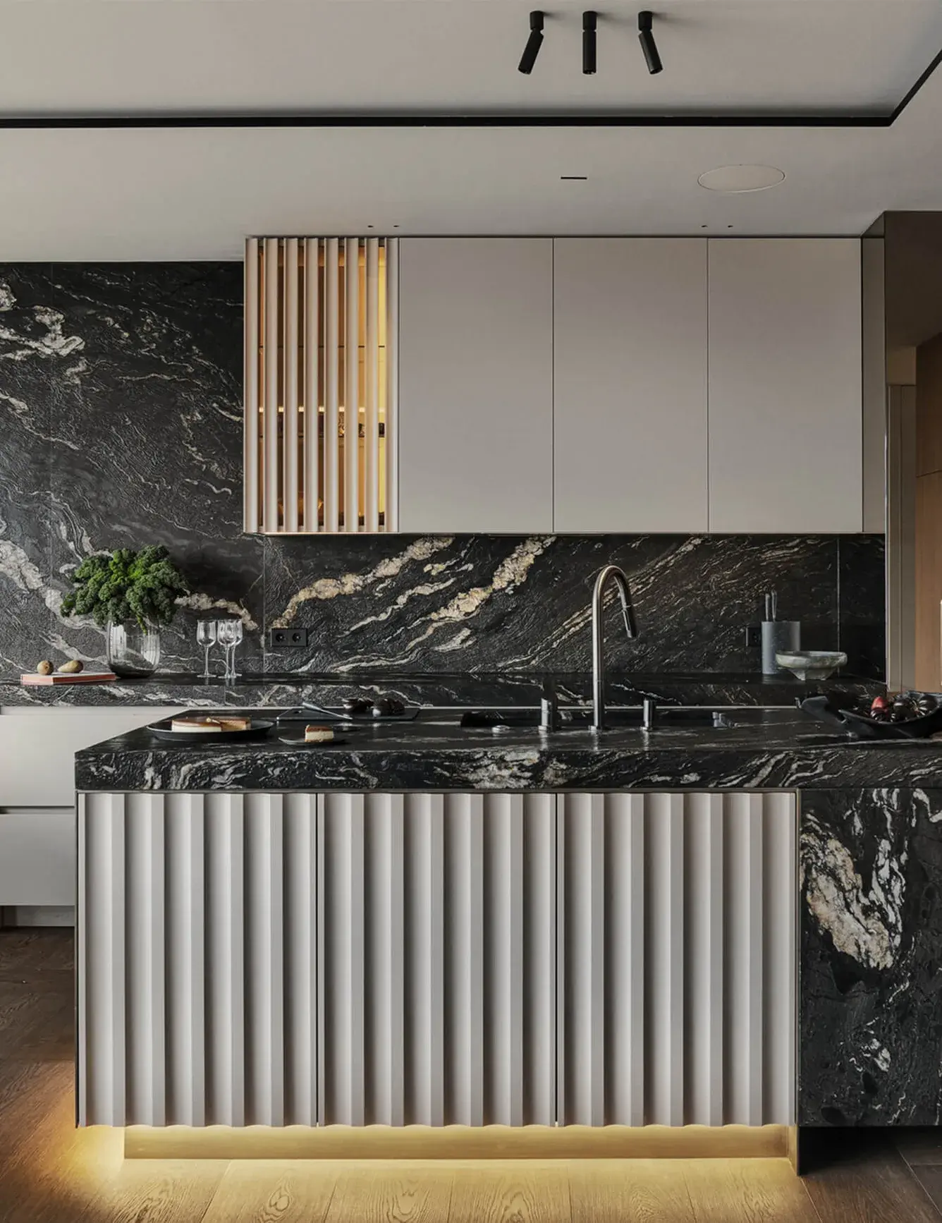 Detailní pohled na luxusní kuchyňský ostrůvek v bytě Grzybowska, který kombinuje tmavý mramor s výrazným žilkováním a čelní panel s vertikálním drážkováním, podsvícený teplým LED páskem u země.