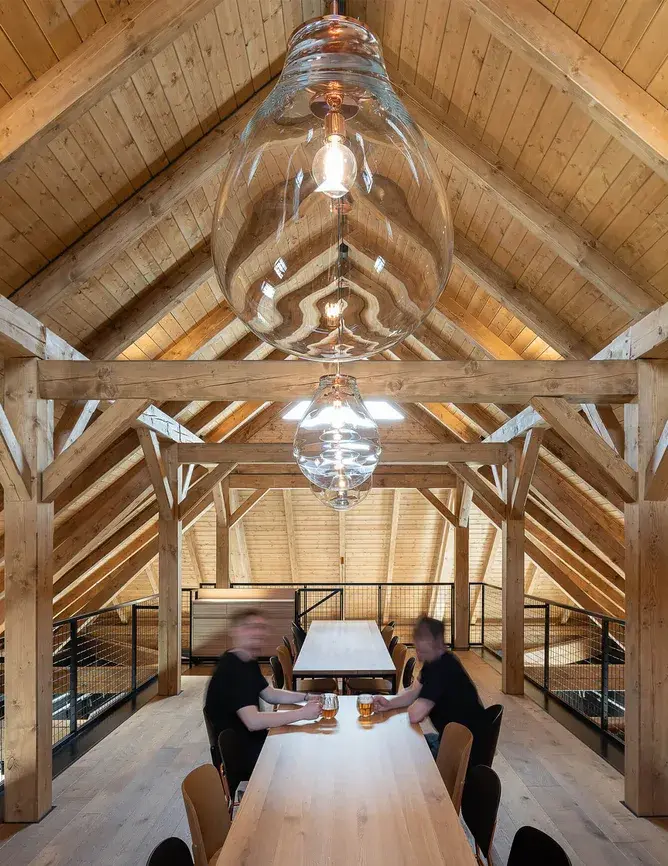 Pohled do podkroví Návštěvnického centra Bernard s odhalenou trámovou konstrukcí krovu, designovými svítidly ve tvaru kapek a dlouhým masivním stolem na světlé dubové podlaze Oakcent.