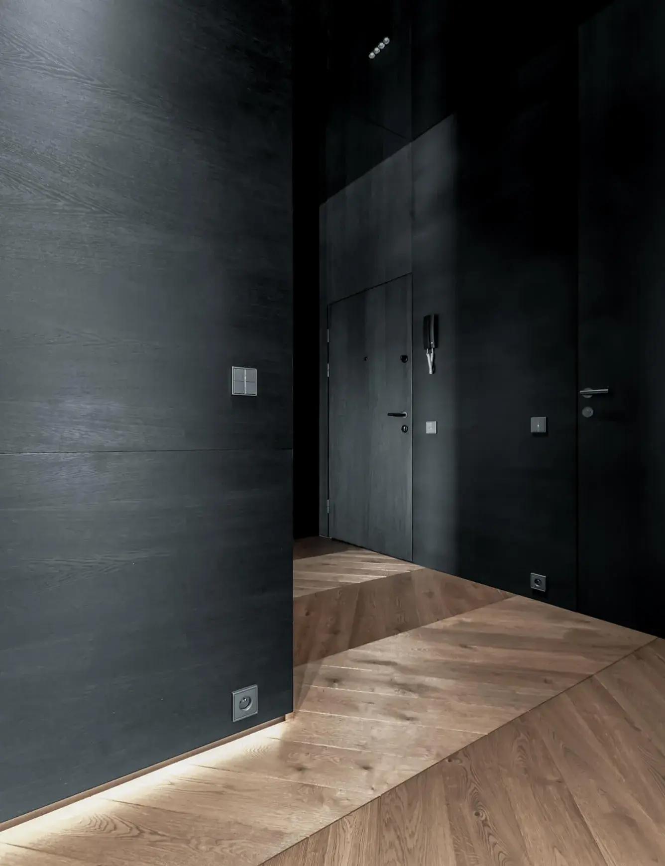 Detailní pohled na moderní chodbu bytu Skierniewicka, kde dubová podlaha Oakcent Rustic ve vzoru Chevron 45 vytváří výrazný geometrický kontrast k minimalistickým černým stěnám a bezfalcovému dveřnímu systému.