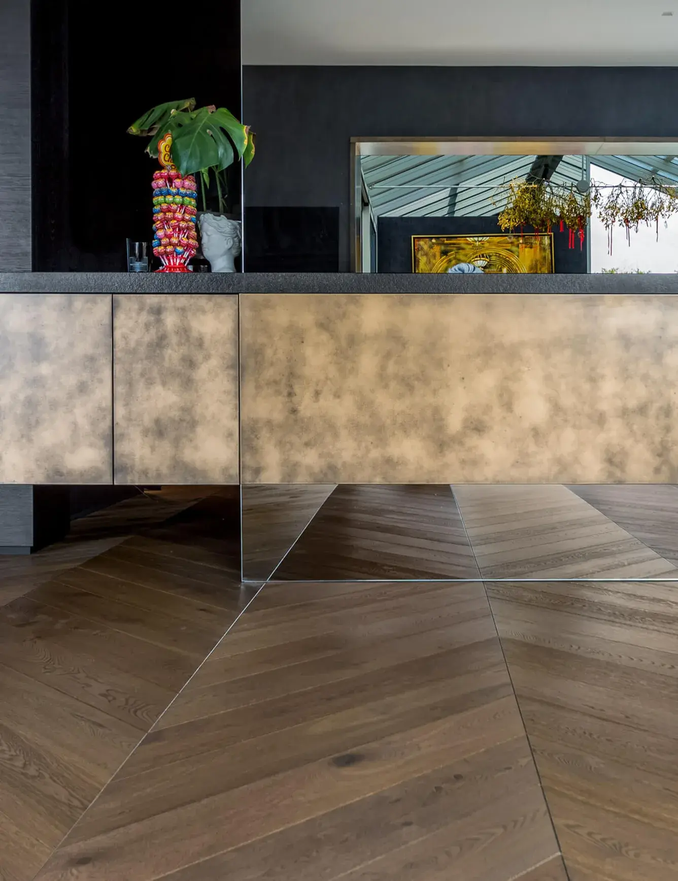 Detailní záběr na kuchyňský ostrůvek s patinovaným povrchem a zrcadlovým soklem v bytě Skierniewicka, pod nímž plynule pokračuje dubová podlaha Oakcent Rustic ve vzoru Chevron 45.