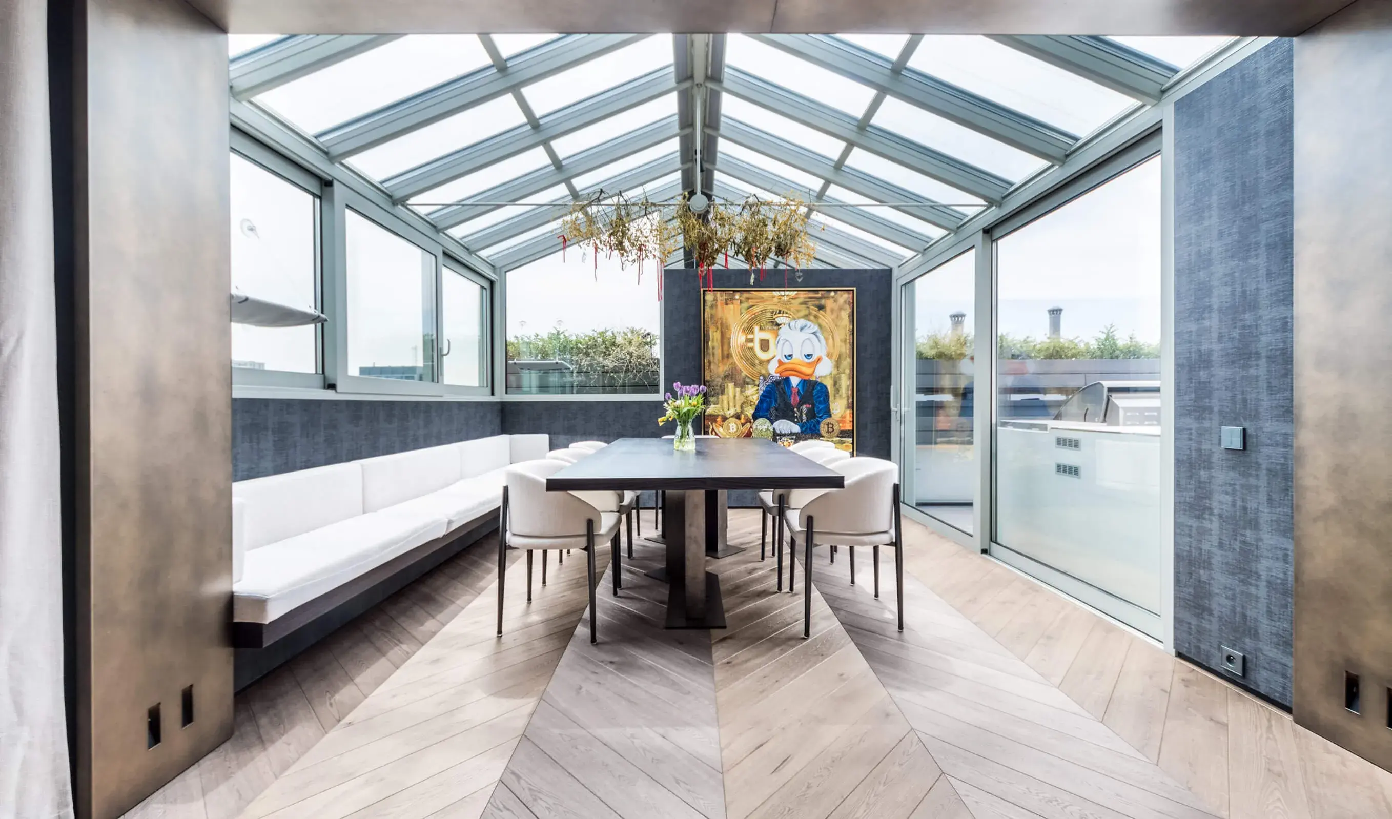 Designová zimní zahrada v varšavském bytě Skierniewicka s velkým jídelním stolem, kde se třívrstvá dubová podlaha Oakcent Rustic v odstínu Grey a vzoru Chevron 45 plynule propojuje s proskleným exteriérem.