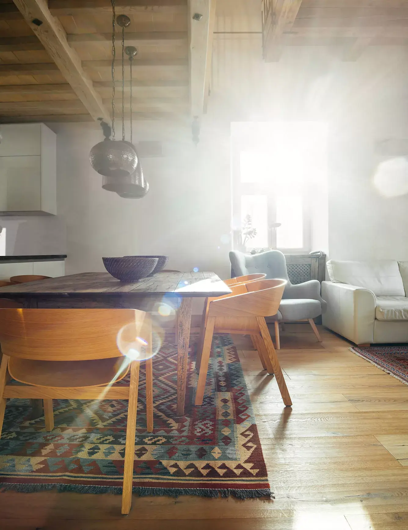 Prosvětlený rustikální interiér v Kaštieli Kubínyi s masivní dubovou podlahou Oakcent Rustic a přiznaným trámovým stropem, kde sluneční paprsky zdůrazňují strukturu dřeva a doplňují historickou atmosféru místnosti s jídelním koutem a vzorovaným kobercem.