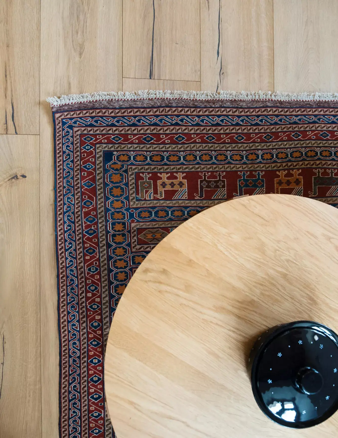 Detailní pohled z nadhledu na část kulatého dřevěného stolku a orientální koberec s bohatým vzorem, položený na přírodní dubové podlaze Oakcent Rustic s výraznou kresbou dřeva.