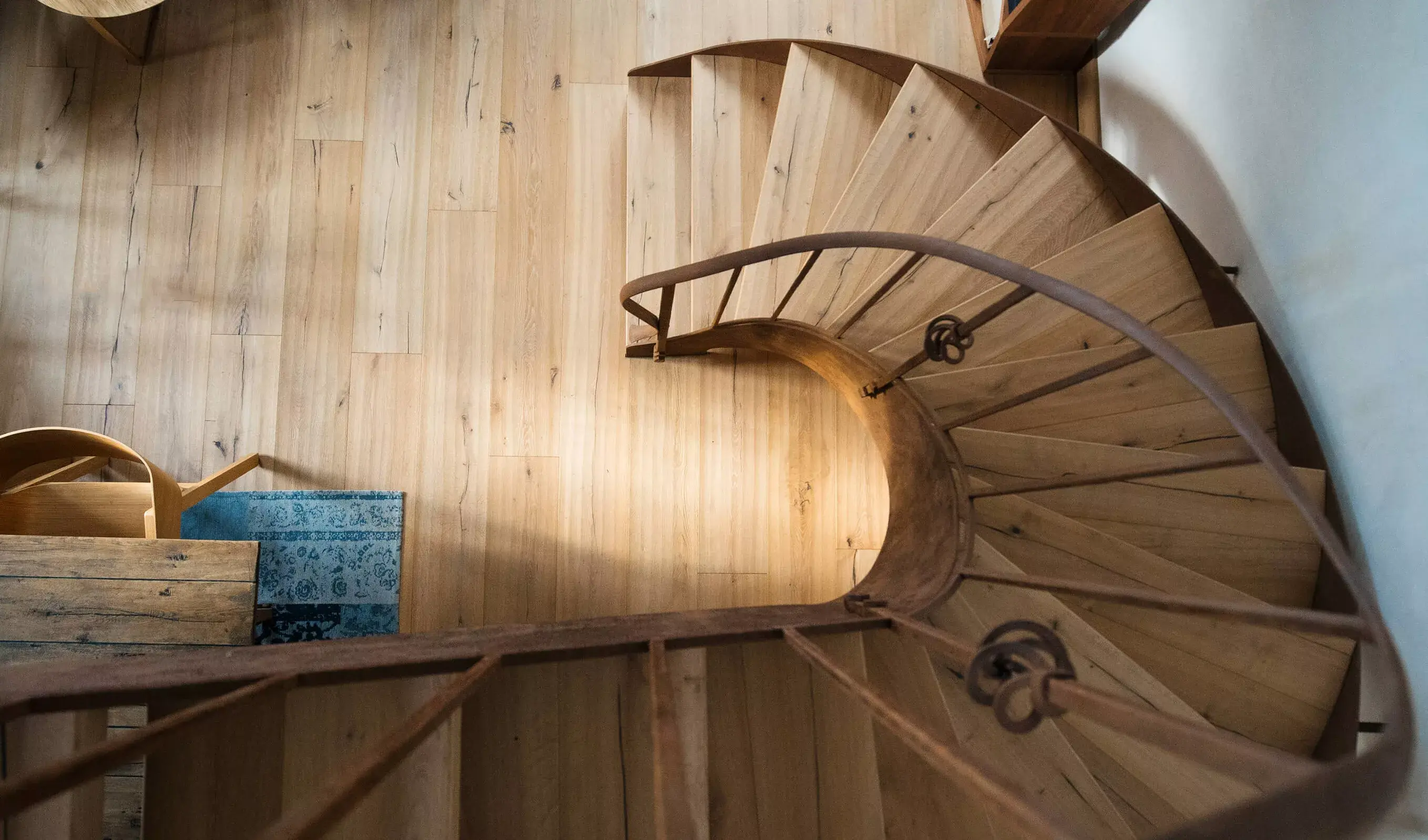 Pohled z nadhledu na točité kovové schodiště s nášlapy ze světlého dubu v historickém interiéru, kde ručně hoblovaná podlaha z kolekce Sand podtrhuje řemeslný charakter celého prostoru.