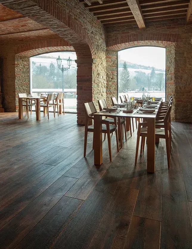 Široký pohled do historické hodovní síně v Kaštieli Kubínyi s tmavou dubovou podlahou Oakcent Rustic, kamennými klenbami a původním trámovým stropem, kde prostřené stoly nabízejí výhled velkými okny do zasněžené zimní krajiny.