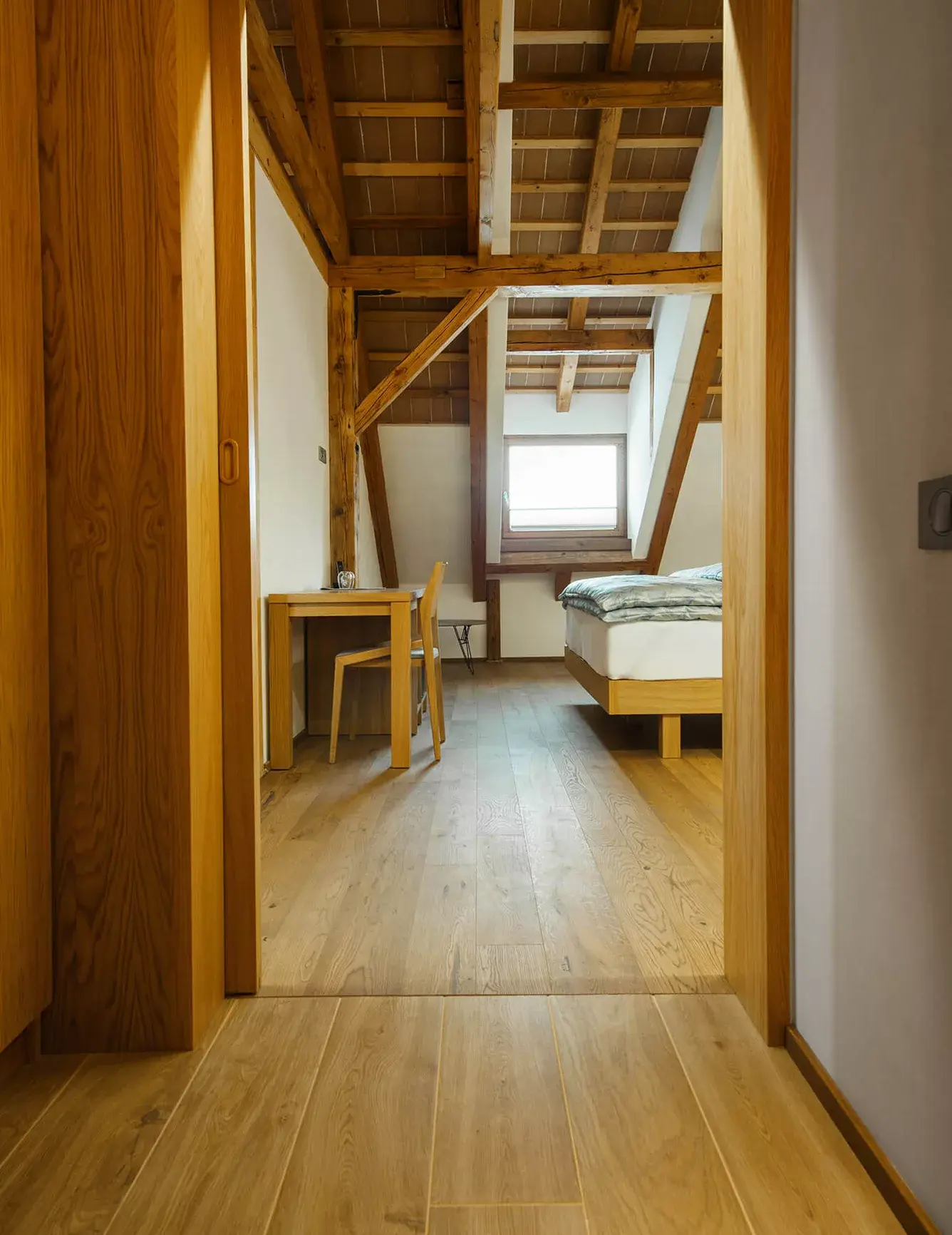 Pohled do útulné podkrovní ložnice v Kaštieli Kubínyi s přiznanou trámovou konstrukcí krovu a světlou dubovou podlahou Oakcent Rustic, která v kombinaci s minimalistickým dřevěným nábytkem vytváří hřejivou atmosféru historického ubytování.