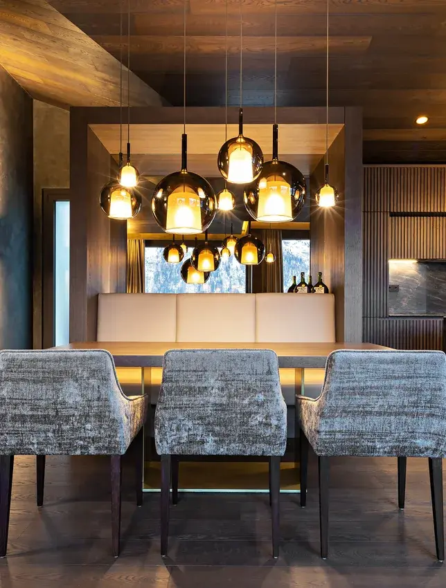 Detailní záběr na luxusní jídelní kout v Rezidenci ve Svatém Mořici se třemi čalouněnými křesly u masivního dřevěného stolu, kterému dominuje skupina designových skleněných svítidel s jantarovým nádechem a kouřová dubová podlaha.