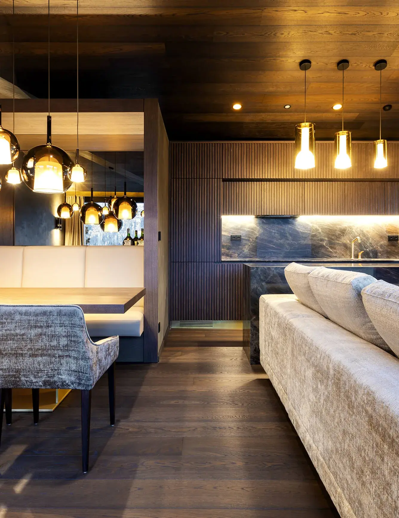 Pohled do moderní kuchyně Rezidence ve Svatém Mořici s tmavým lamelovým obložením stěn a integrovanou kuchyňskou linkou s mramorovou pracovní deskou, kterou doplňuje tmavá dřevěná podlaha a designové visací osvětlení nad jídelní částí.